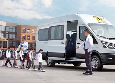 Students boarding a white Ford Transit school bus with a driver and teacher in front of a school building.