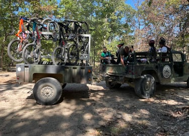 Military vehicle towing a bike rack trailer loaded with mountain bikes for riders on a wooded trail.