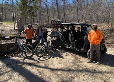 A group of mountain bikers with their gear and a black shuttle utility vehicle on a wooded trail.