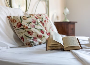 An open hardcover book rests on a white bed with floral pillows in a cozy bedroom setting.