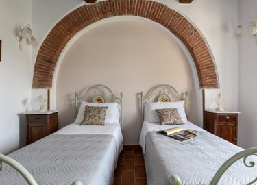 Rustic twin bedroom with ornate metal headboards under a large brick archway in an Italian villa.