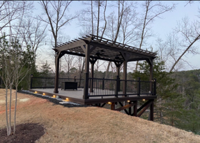 Elevated wooden deck with a dark pergola and black metal railing overlooking a forest at dusk.