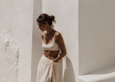 A woman in a cream linen sarong and crop top leans against a white textured Mediterranean wall.
