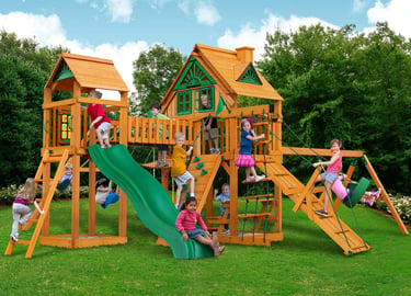 Children playing on a large cedar wood backyard swing set with a slide and playhouse.