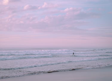 Serene shot of man in the distance standing in the ocean's waves. Pastel sunset sky.