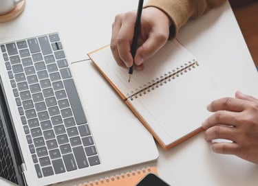 someone writing with a pen in a note book a laptop computer is on the desk