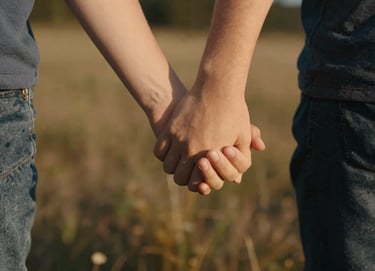 A close-up candid of a child's hand holding a parent's hand, softly lit by the warm afternoon sun. The background is a soft-focus meadow of earthy warm brown and deep charcoal shadows.