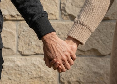 A refined, close-up shot of two people holding hands against a soft sand colored stone wall. One person wears a charcoal sleeve, the other a warm beige knit. The lighting is natural and directional, highlighting the textures and the authentic connection. Elegant and storytelling-focused.