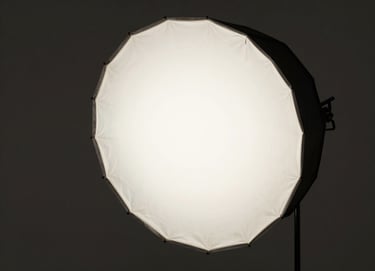 A side view of a large studio softbox diffusing light in a dark, minimalist space. The atmosphere is professional and quiet, reflecting a North American artistic workshop aesthetic. Colors are restricted to black, dark gray, and off-white.