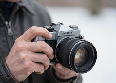 A close-up, detailed shot of a photographer's hand adjusting a vintage camera lens. The person is wearing a dark charcoal leather jacket in a Global / Western outdoor setting. The focus is sharp on the mechanical details of the camera with a blurred soft white background.