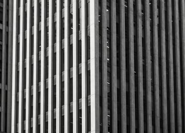 A vertical architectural photograph of a modern building facade featuring clean vertical lines and glass. The image is strictly monochrome with deep blacks and bright whites. Sharp shadows create a geometric pattern. North American / European city center.