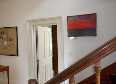 Oil painting of a sunset on a gallery wall near a wooden staircase and white doorway.