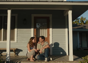 A couple sharing a private, authentic laugh while sitting on the porch of a classic North American / US home. The scene is shot in a cinematic style with warm, sun-drenched lighting and deep charcoal shadows for contrast.
