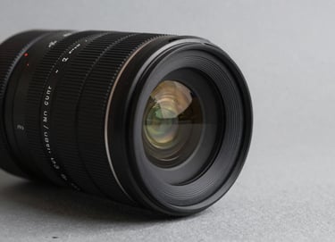 Detail shot of a professional camera lens on a soft gray surface, sharp focus, minimal composition, charcoal black accents, Global / Contemporary Art.