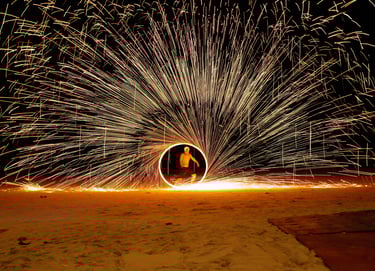 A fire spinner performs on a beach at night, creating a circle of sparks.- by ACAT PHOTOS