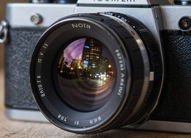 A close-up of a vintage 35mm film camera lens reflecting the lights of a North American / US city at night. The image has a polished, high-end feel with steel gray metallic textures and deep onyx shadows.