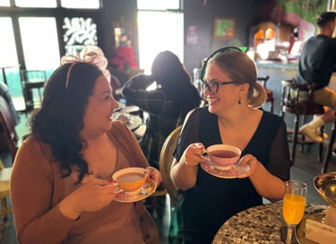 Two women wearing small headpieces are holding tea cups and smiling at each other.