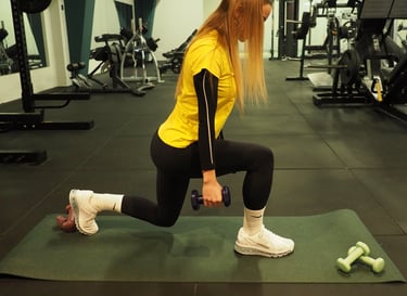 Una donna in maglia gialla esegue un affondo con manubri in una palestra ben attrezzata, concentrandosi su una corretta postu