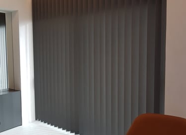 Modern charcoal grey vertical blinds installed on a large window with warm cove lighting.