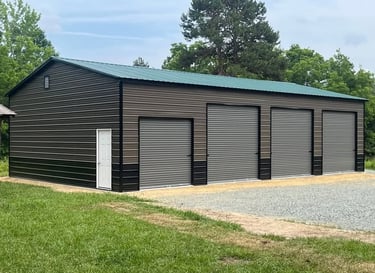 Large brown metal garage building with a green roof and four roll-up doors on a gravel pad.