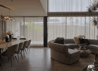 Sala-comedor con cortinas traslúcidas de doble altura, sofá en escuadra y mesa de mármol | KARÄNA
