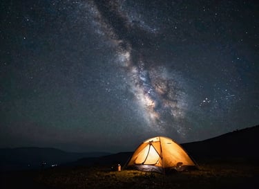 Wide starscape with the Milky Way arching over a quiet mountain camp, a single silhouetted tent glowing with warm amber light inside.