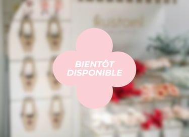Pink flower-shaped 'Bientôt Disponible' coming soon sign overlaying a blurred boutique flower shop interior.