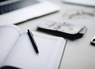 A black pen resting on an open paper planner next to a stack of cash and a laptop on a white desk.
