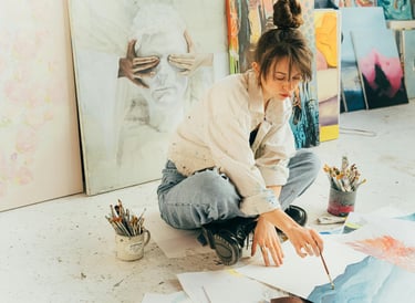 Jovem artista sentada no chão rodeada por pinturas, trabalhando em uma aquarela.