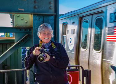 A fotógrafa Martha Cooper, segurando uma câmera e sorrindo, posa em um ambiente urbano