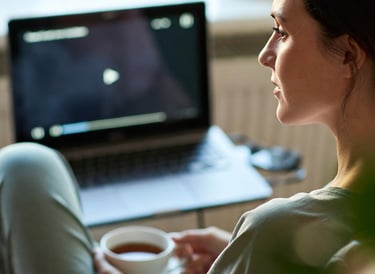 A woman relaxing at home watching a video on her laptop while holding a cup of hot tea.