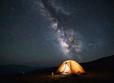 Wide starscape with the Milky Way arching over a quiet mountain camp, a single silhouetted tent glowing with warm amber light inside.