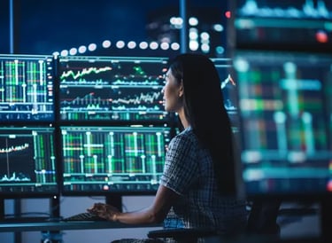 Female financial analyst monitoring stock market data and real-time trading charts on multiple computer monitors.