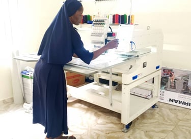 A nun in blue habit operating a white professional multi-needle embroidery machine in a workshop.