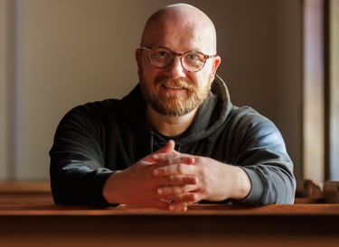 A man with glasses sitting in a church pew, smiling at the camera