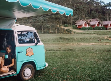 Uma pessoa sentada bebendo café dentro de uma kombi home, estacionada em um jardim.