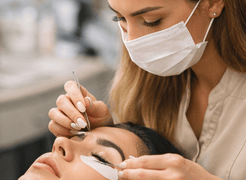 Professional lash artist applying eyelash extensions to a client using tweezers under a ring light.