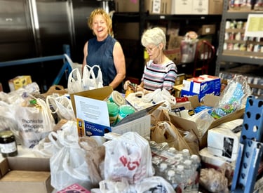 Two women standing behind table covered in bags of donated food in food pantry