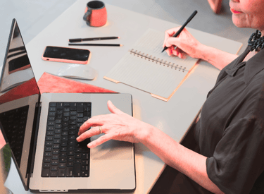 Uma mulher consulta o notebook aberto em cima da mesa enquanto faz anotações no caderno ao lado.