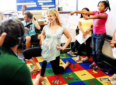 Group of children in a classroom move around a smiling woman kneeling