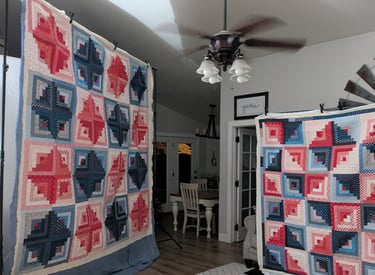 Two red white and blue colorful log cabin quilts on display stands in a cozy room with wood floors.
