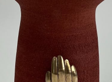 A tall thrown red ceramic vessel, wide at the top. A cast bronze hand is fixed on its base.