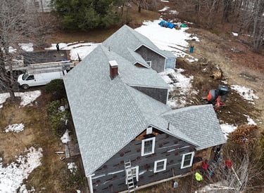 Aerial view of a residential roof replacement and home renovation project with grey shingles and excavating equipment.