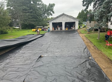 Workers laying black geotextile fabric for driveway stabilization in front of a residential garage.