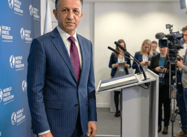 A speaker in a suit delivering remarks at an international press event, standing behind a podium. 