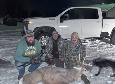 Three hunters posing in the snow with a harvested buck and thermal drone near a white truck.