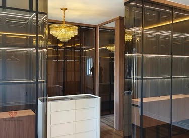 Modern luxury walk-in closet with gold crystal chandelier, LED-lit glass shelving, and white island dresser.