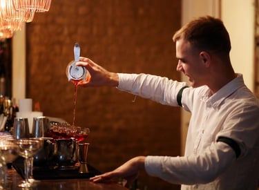 Professional bartender straining a red craft cocktail into glasses at a dimly lit upscale bar.