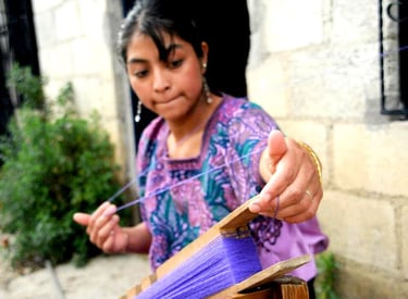 A skilled artisan weaver in Guatemala handcrafting textiles with purple yarn on a traditional loom.