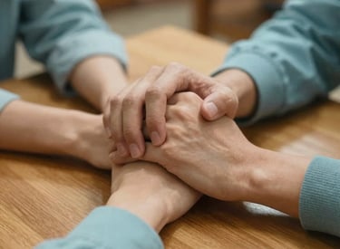 A close-up photograph of two people's hands clasped together across a wooden table, conveying trust and support. The setting is a warm North American community center. Soft Honeydew and Celadon Blue tones are present in the lighting.